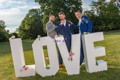 Trois hommes en costume posent devant de grandes lettres formant le mot ’LOVE’ dans un parc verdoyant.
