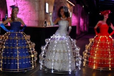 Trois femmes en robes lumineuses portant des structures en verre à pied, dans un espace éclairé aux tons violets.