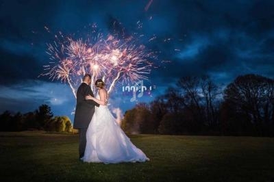 Un couple en tenue de mariage regarde un feu d'artifice illuminant un ciel nocturne nuageux dans un parc.