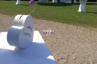 Urne blanche en forme de cœur et livre d'or sur une table, avec des ballons et une réception en plein air en arrière-plan.