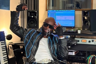 Un homme assis dans un studio d'enregistrement, portant une tenue noire avec des motifs lumineux, devant du matériel audio.