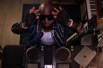 Un DJ portant des lunettes de soleil ajuste sa tenue devant une table de mixage dans un studio équipé d'instruments.