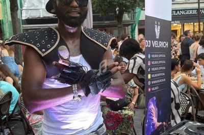 Un DJ en tenue originale pose devant ses platines dans une rue animée avec des terrasses de café en arrière-plan.