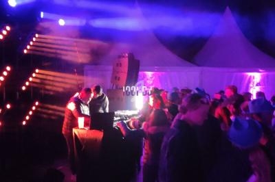 Des personnes dansent devant un DJ sous des lumières colorées lors d'un événement nocturne en extérieur.