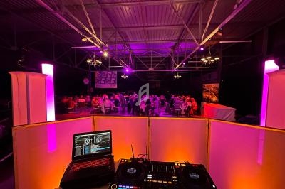 Vue depuis une cabine de DJ avec platines et ordinateur, éclairage coloré et invités assis dans une salle sombre.