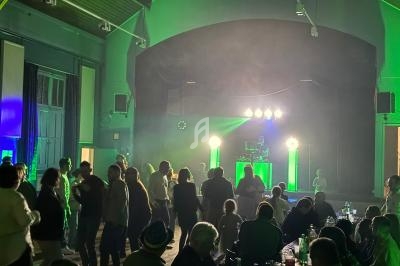 Des personnes dansent et discutent dans une salle éclairée par des lumières vertes, avec un DJ sur une scène au fond.