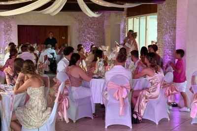 Salle de réception décorée avec des nappes blanches et roses, des invités assis autour de tables lors d'un événement festif.