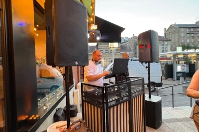 Un DJ mixe en extérieur devant un restaurant, entouré de haut-parleurs, au crépuscule dans une rue animée.