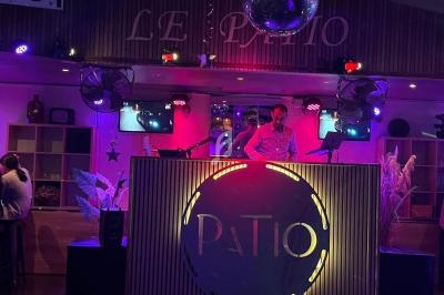 Un DJ mixe sur une scène éclairée par des lumières colorées dans un lieu nommé ’Le Patio’.