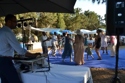 Des personnes dansent sur une piste bleue en extérieur, près d'un DJ, entourées de tentes et d'arbres.