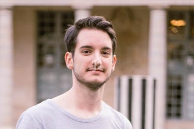 Jeune homme souriant devant un bâtiment avec des colonnes et des fenêtres en arrière-plan.