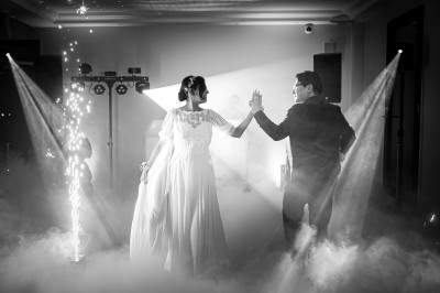 Un couple en tenue de mariage danse au milieu de la fumée et des lumières dans une salle sombre.