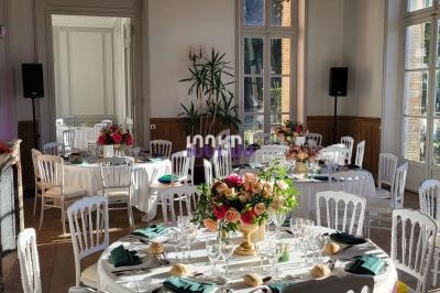 Salle de réception lumineuse avec tables rondes dressées, décorées de fleurs et entourées de chaises blanches.