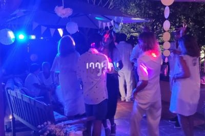 Des personnes en tenue blanche dansent et discutent lors d'une soirée en extérieur, éclairée par des lumières colorées.