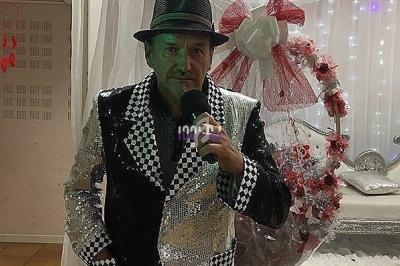 Homme en costume brillant noir et argent avec un chapeau, se prenant en photo devant une décoration florale blanche et rouge.