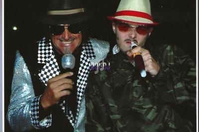 Deux hommes chantant au micro, portant des chapeaux et des tenues originales, dans un environnement sombre.