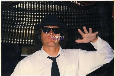 Homme portant un chapeau noir, des lunettes de soleil et une chemise blanche, levant une main devant lui.