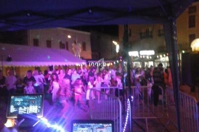 Vue nocturne d'une fête en plein air avec une foule dansante, des lumières colorées et des équipements de DJ au premier plan.