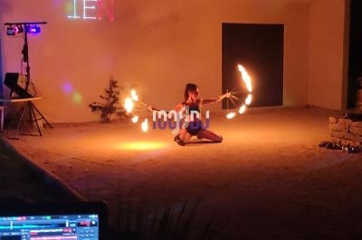 Une artiste jongle avec des bâtons enflammés dans une cour extérieure éclairée par des lumières colorées.