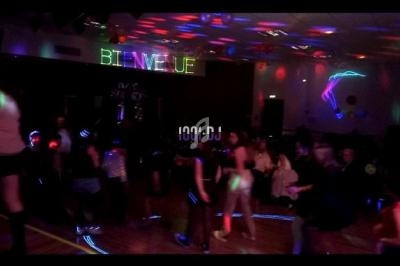 Salle sombre avec des jeux de lumière colorés, des personnes dansant et un panneau lumineux affichant ’Bienvenue’.