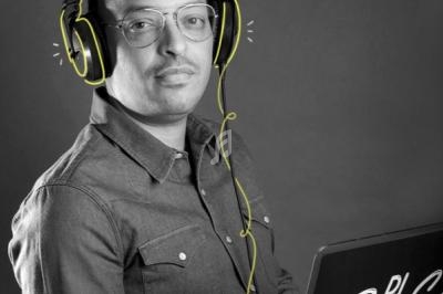 Homme portant un casque audio et des lunettes, assis devant un ordinateur portable, sur fond gris.