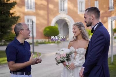 Un couple de mariés discute avec un homme devant un bâtiment en briques rouges et un jardin.
