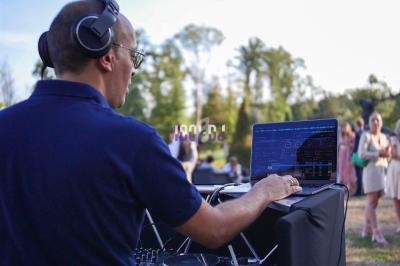 Un DJ mixe de la musique en extérieur, avec un ordinateur portable et une table de mixage, devant un public en arrière-plan.