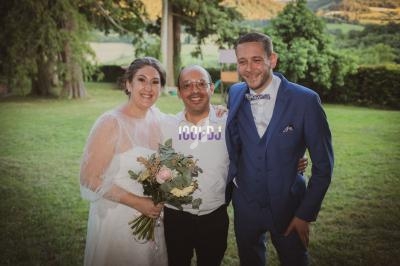 Un couple en tenue de mariage pose avec un homme souriant dans un jardin verdoyant au coucher du soleil.