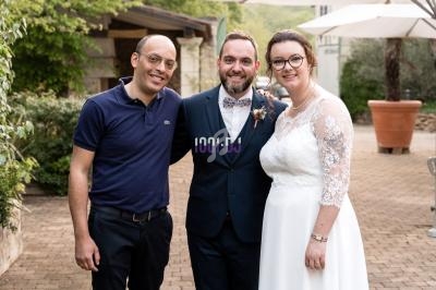 Un homme pose avec un couple en tenue de mariage dans une cour extérieure pavée et verdoyante.