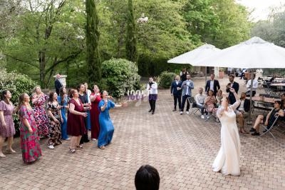 Un groupe de personnes assiste à un lancer de bouquet par une mariée dans une cour pavée entourée de verdure.