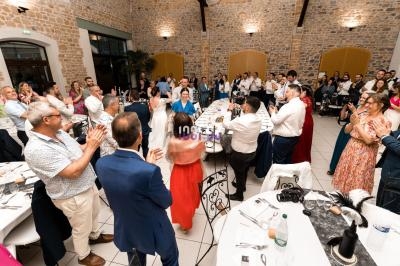 Des invités debout applaudissent autour de tables dressées dans une grande salle en pierre lors d'un événement festif.