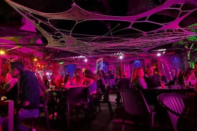 Des personnes assises à des tables dans un bar ou restaurant en extérieur, éclairé par des lumières colorées et décoré de…