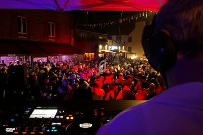 Un DJ mixe sous une tente lors d'une fête nocturne en plein air, avec une foule animée et des lumières colorées.