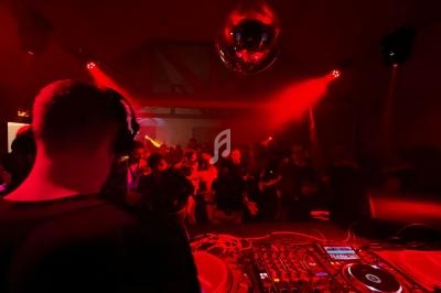 Un DJ mixe sur une scène éclairée de lumières rouges, face à une foule dansante sous une boule disco.