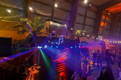 Un DJ mixe sur une scène décorée de plantes sous des lumières colorées, entouré de spectateurs dans une salle.