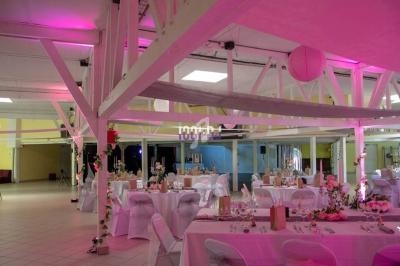 Salle de réception décorée avec des tables rondes dressées, éclairage rose et poutres apparentes blanches.