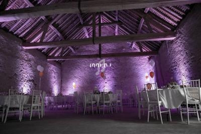 Salle en pierre avec charpente apparente, décorée de lumières violettes, tables dressées et ballons colorés.