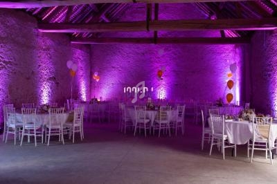 Salle de réception en pierre avec tables décorées, chaises blanches et éclairage violet, ballons suspendus.