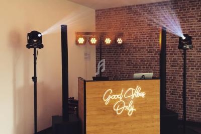 Poste de DJ avec une table en bois éclairée par un néon ’Good Vibes Only’, entourée de projecteurs et d'enceintes.