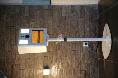 Borne interactive blanche avec écran tactile et appareil photo, installée devant un mur texturé gris.