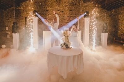 Un couple de mariés célèbre devant un gâteau de mariage, entouré de fumée, lumières et étincelles dans une salle en pierre.