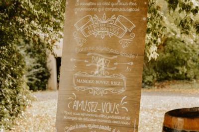 Panneau décoratif de mariage en extérieur, avec inscriptions manuscrites et motifs floraux, posé sous un arbre.
