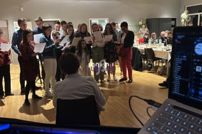 Un groupe de personnes chante debout dans une salle éclairée, avec un public assis à des tables en arrière-plan.