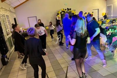 Des personnes dansent et discutent dans une salle décorée de ballons noirs et jaunes, éclairée par des lumières festives.