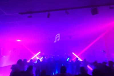 Des silhouettes dansent sous des lumières violettes et roses dans une ambiance festive et enfumée.