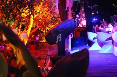 Projecteur éclairant une terrasse extérieure avec des chaises modernes, entourée de végétation et d'éclairage coloré.