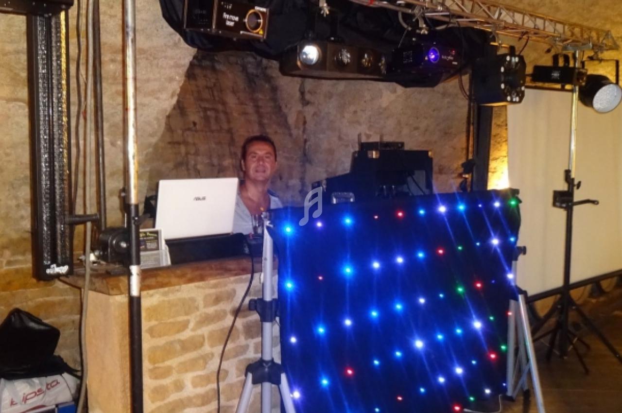 Un DJ derrière une table équipée de matériel audio et d'un panneau lumineux coloré dans une salle en pierre.