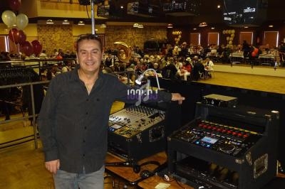 Un homme souriant se tient près d'une table avec du matériel de sonorisation dans une salle remplie de personnes.