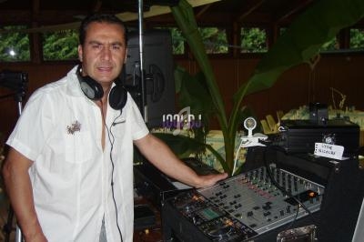 Homme debout près d'une table de mixage, portant un casque audio autour du cou, dans un espace intérieur avec des plantes.