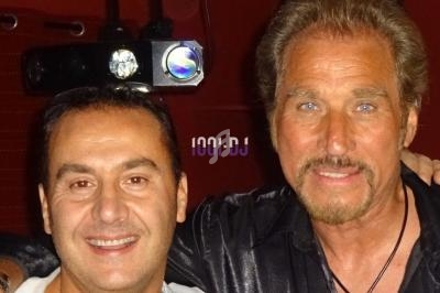 Deux hommes posant ensemble devant un fond rouge, l'un souriant et l'autre avec une expression neutre.